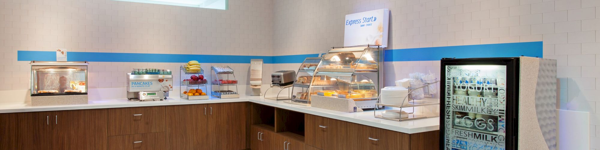 The image shows a clean, modern breakfast buffet area with various food items on display, including a pastry case and a refrigerated section.