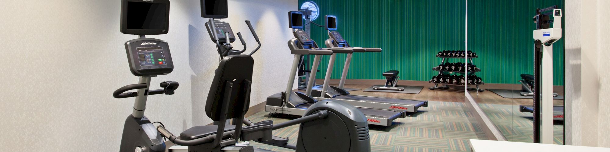 The image shows a gym with exercise equipment, including stationary bikes, treadmills, a set of dumbbells, and a wall-mounted TV on the left side.