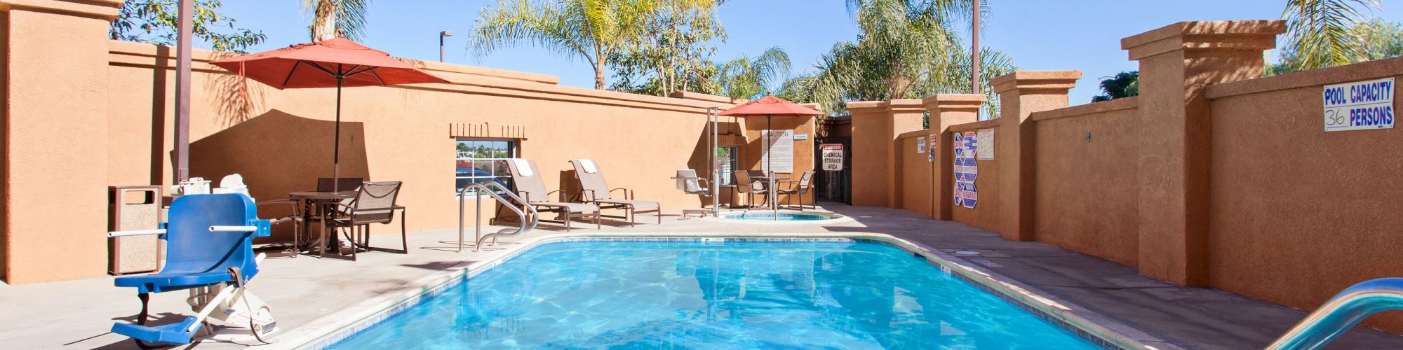 An outdoor swimming pool surrounded by lounge chairs, umbrellas, palm trees, and a lift for accessibility.