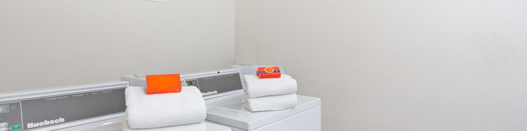 The image shows a laundry room with two washing machines, each topped with neatly folded towels and an orange item on top.