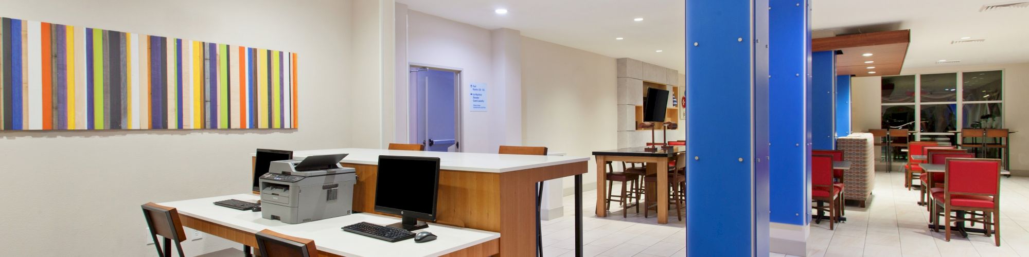 A modern communal workspace with computers on desks; adjacent seating area with red chairs and tables, featuring bright lighting and wall art.