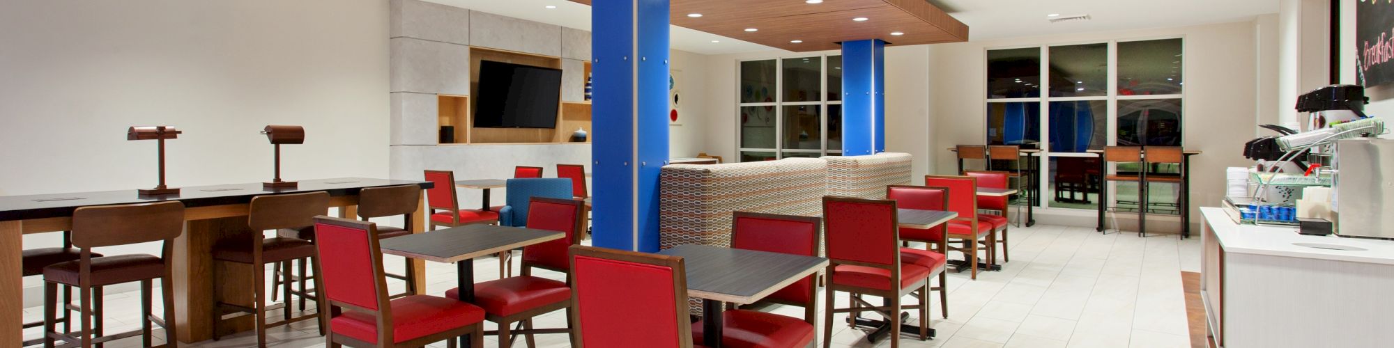 A modern dining area with red chairs, tables, a TV, counter, and coffee machine. Bright lighting and minimalist décor.