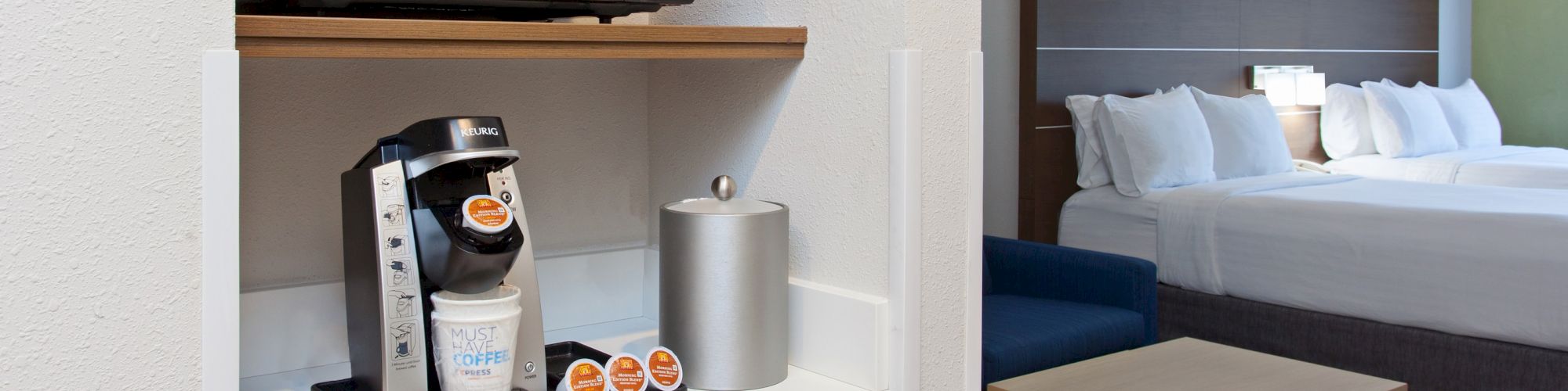 The image shows a hotel room with a microwave, coffee maker, mini-fridge, and two neatly made beds in the background, including a bedside table.