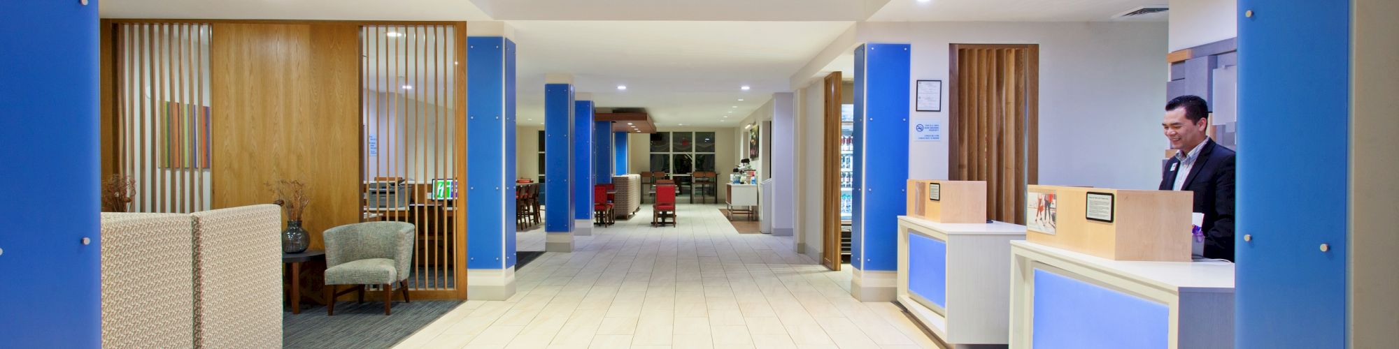 A modern hotel lobby with a reception desk, a person at the counter, seating areas, and bright lighting, with blue accents and wooden elements.