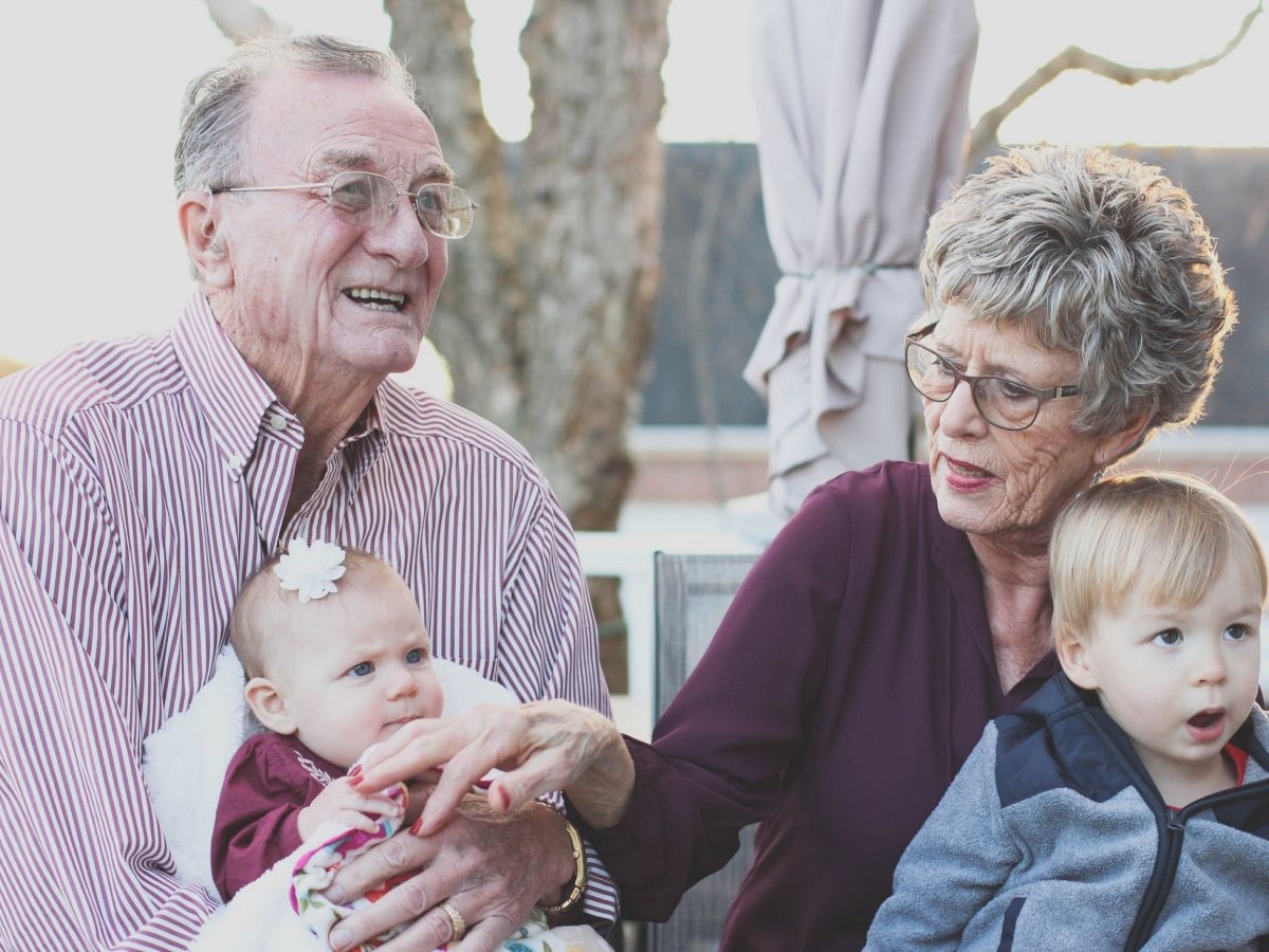 An elderly couple is sitting outside, holding a baby and a toddler, enjoying a moment together in what appears to be a backyard setting.