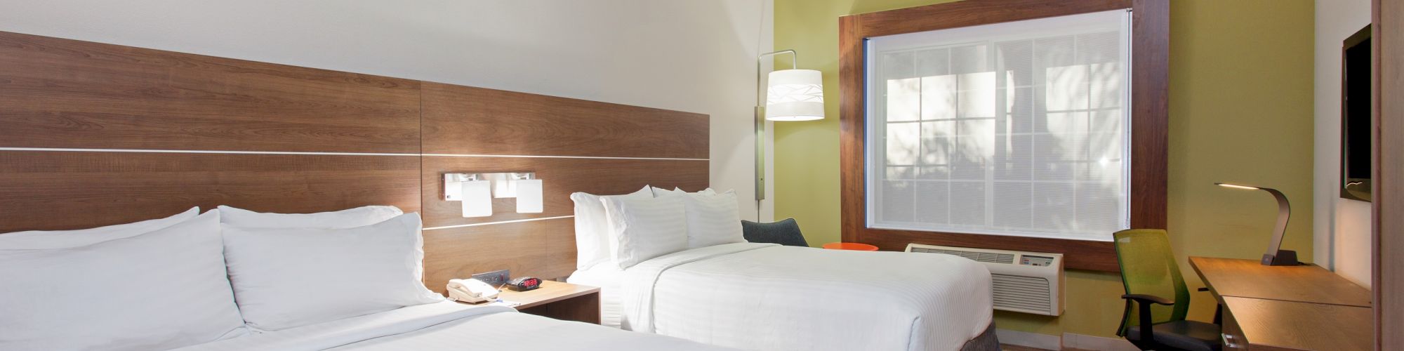 The image shows a tidy hotel room with two double beds, a flat-screen TV, a desk, a chair, and a window with a view, under warm lighting.