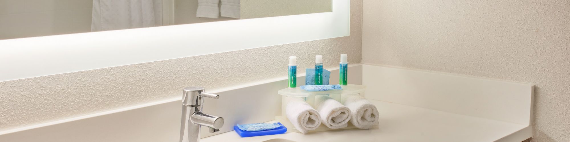 A bathroom countertop with a sink, toiletries, folded towels, a toothbrush, and a large illuminated mirror is pictured.