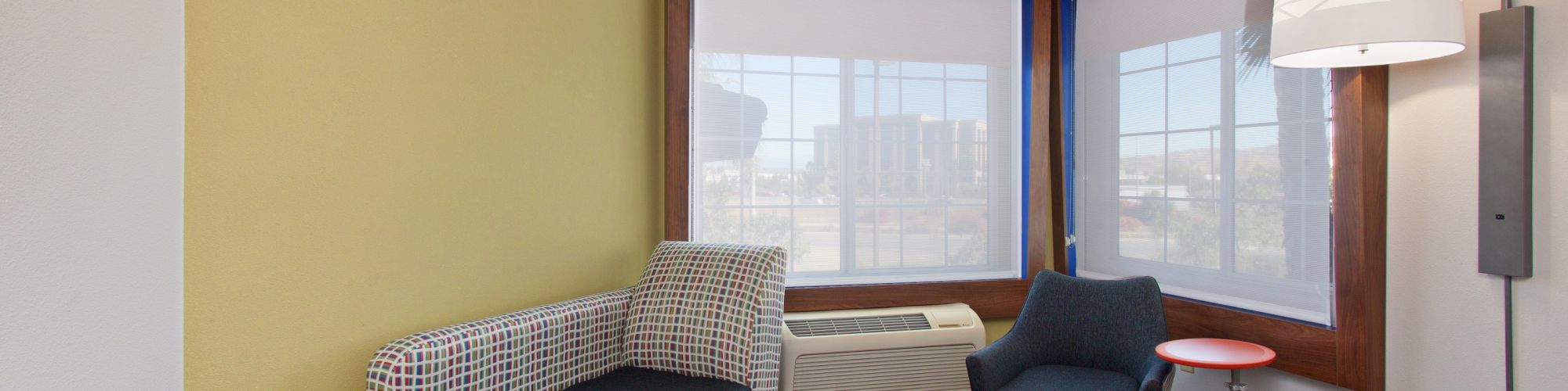 A modern hotel room features a sofa, an armchair, a side table, large windows, and a floor lamp, with light wooden flooring and a yellow accent wall.