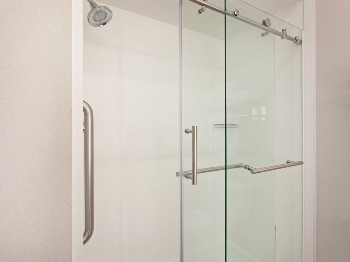 The image shows a modern shower with a sliding glass door, metal handles, and a white interior.