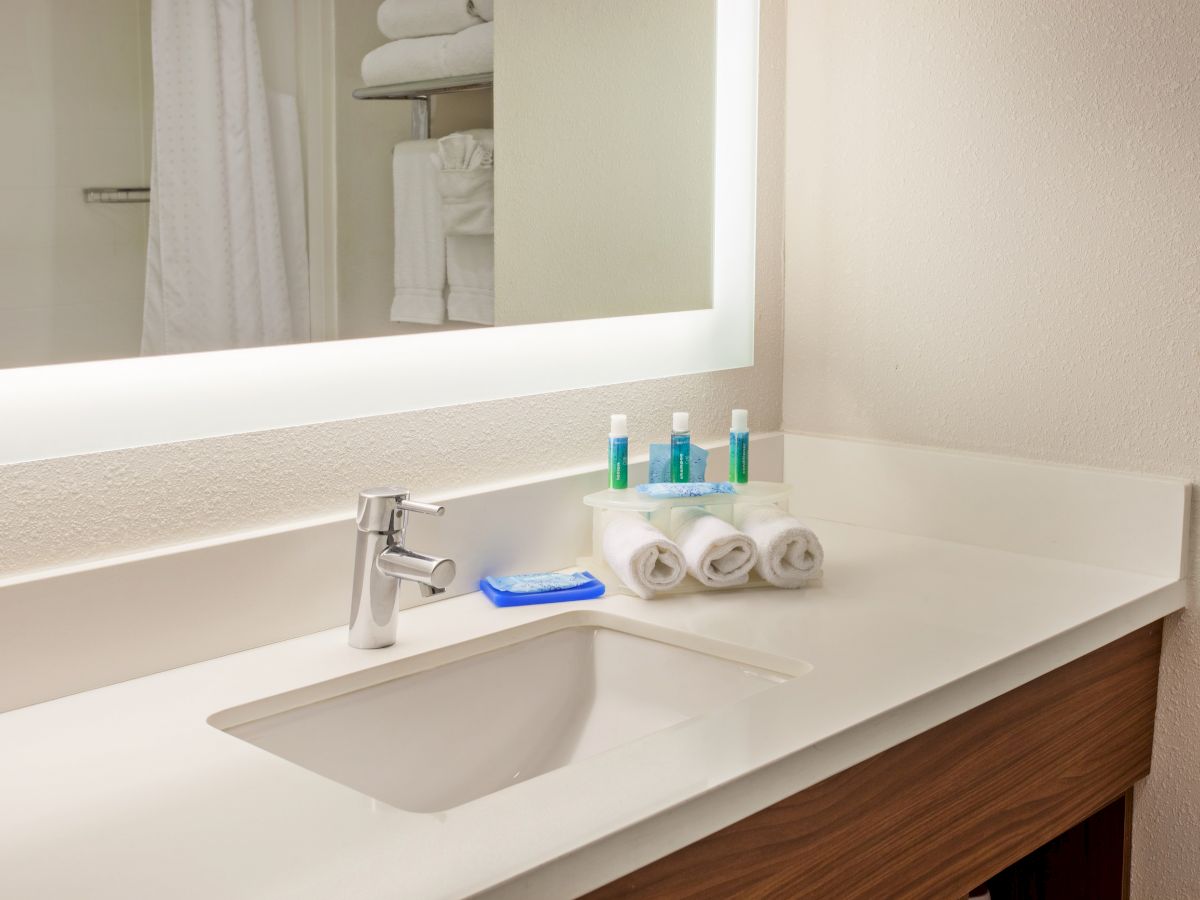 A clean bathroom countertop with a sink, a faucet, rolled towels, toiletries, and a large illuminated mirror in the background.