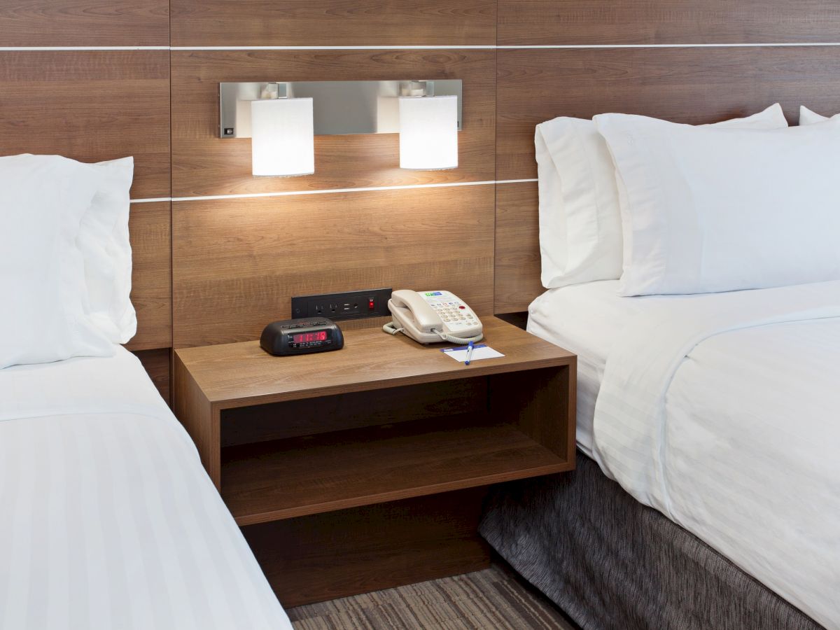 A hotel room scene shows two beds with white linens, a wooden nightstand in between, and a lamp, telephone, and alarm clock on the nightstand.