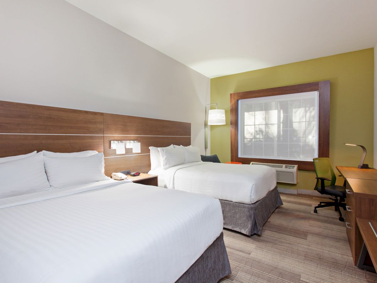 The image shows a modern hotel room with two beds, wooden furniture, a desk with a chair, and a large window with a green accent wall.