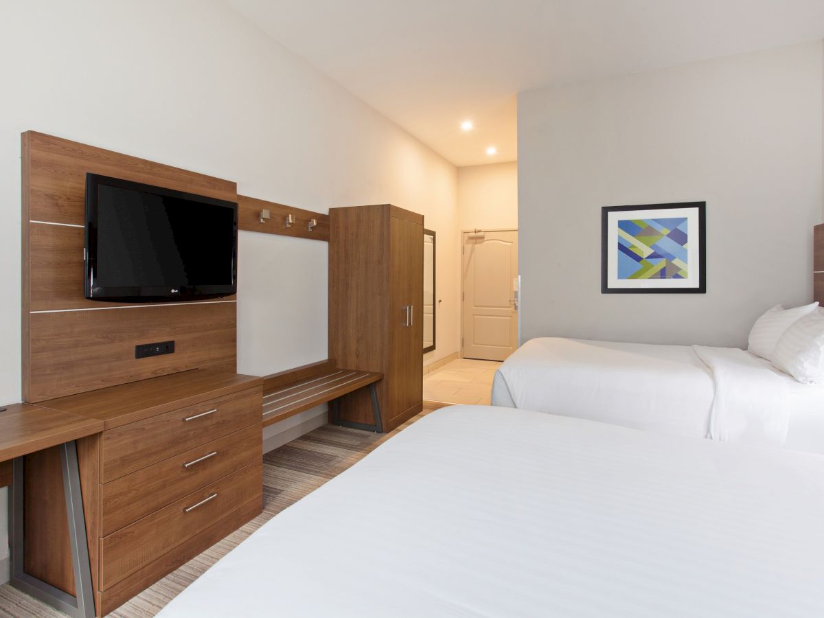 A hotel room with two beds, a wall-mounted TV, a desk, a closet, and a framed picture on the wall, featuring modern decor and lighting.