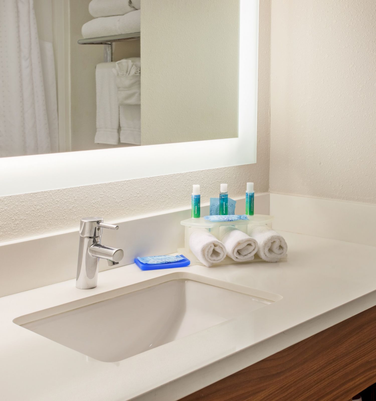 A bathroom countertop with a sink, faucet, soap bar, rolled towels, and various toiletries is placed in front of a large mirror.