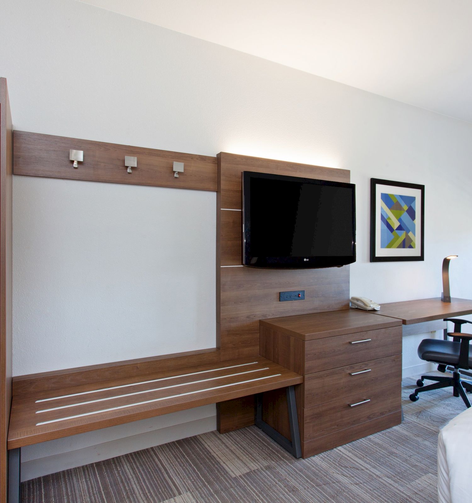 This image shows a hotel room workstation with a wall-mounted TV, wooden desk, chair, artwork, and a wardrobe next to a balcony door.