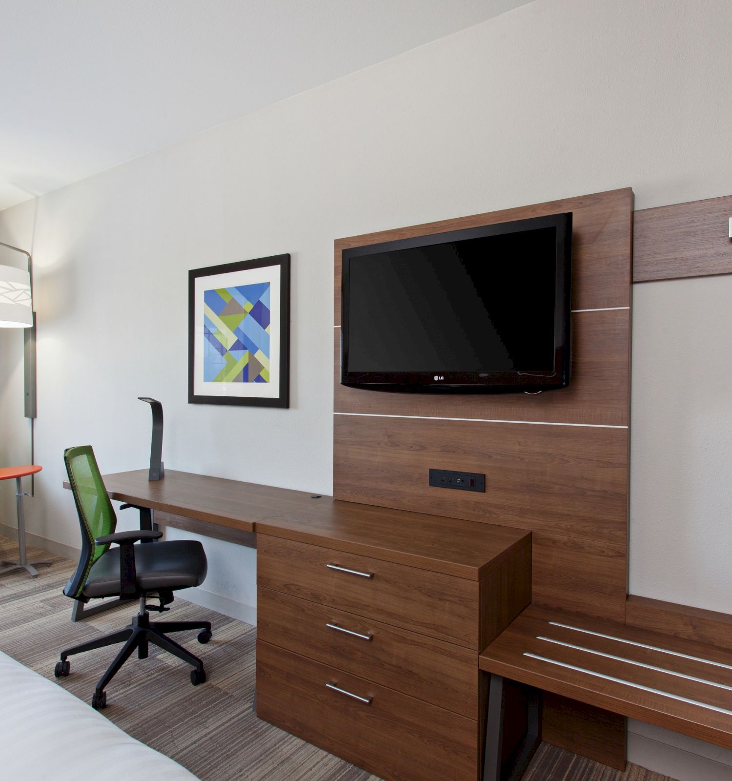 The image shows a modern hotel room with a desk, chair, flat-screen TV, wall art, bench, wall-mounted coat hooks, single bed, armchair, and floor lamp.