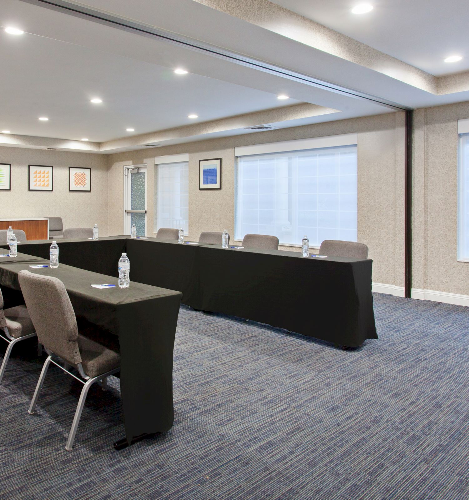 The image shows a conference room with U-shaped table arrangement, chairs, bottled water, and framed artwork on the walls, under ceiling lights.