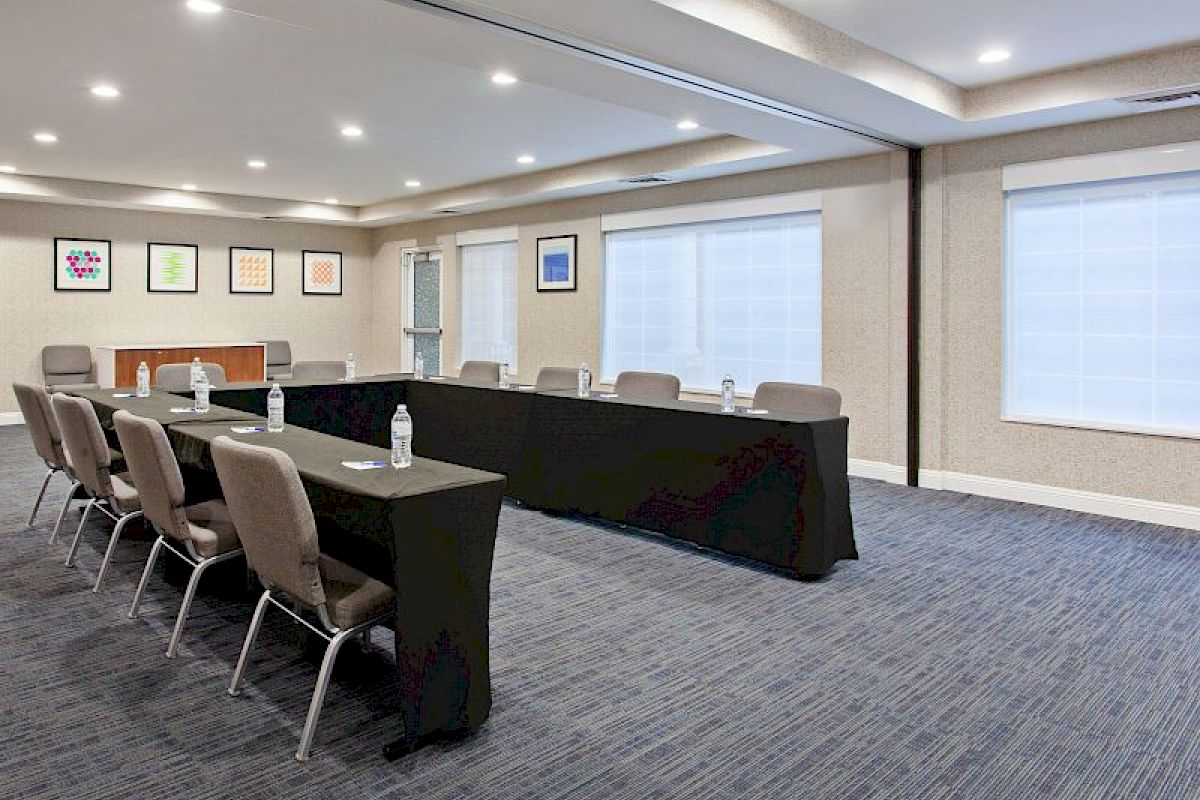 This image shows a conference room with a U-shaped table setup, chairs, water bottles, framed art on walls, and large windows with blinds.