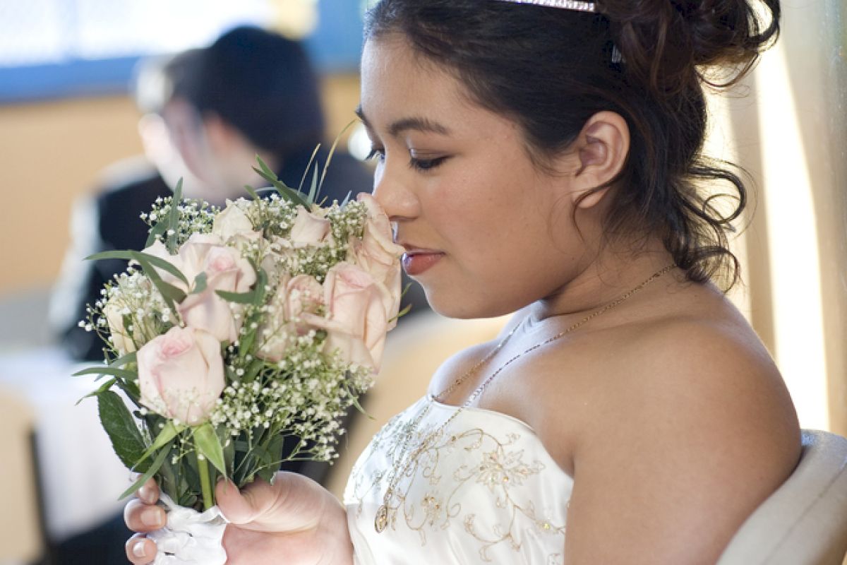 A bride in a white dress and tiara sits on a chair, holding and sniffing a bouquet of pink roses, with a blurred background.