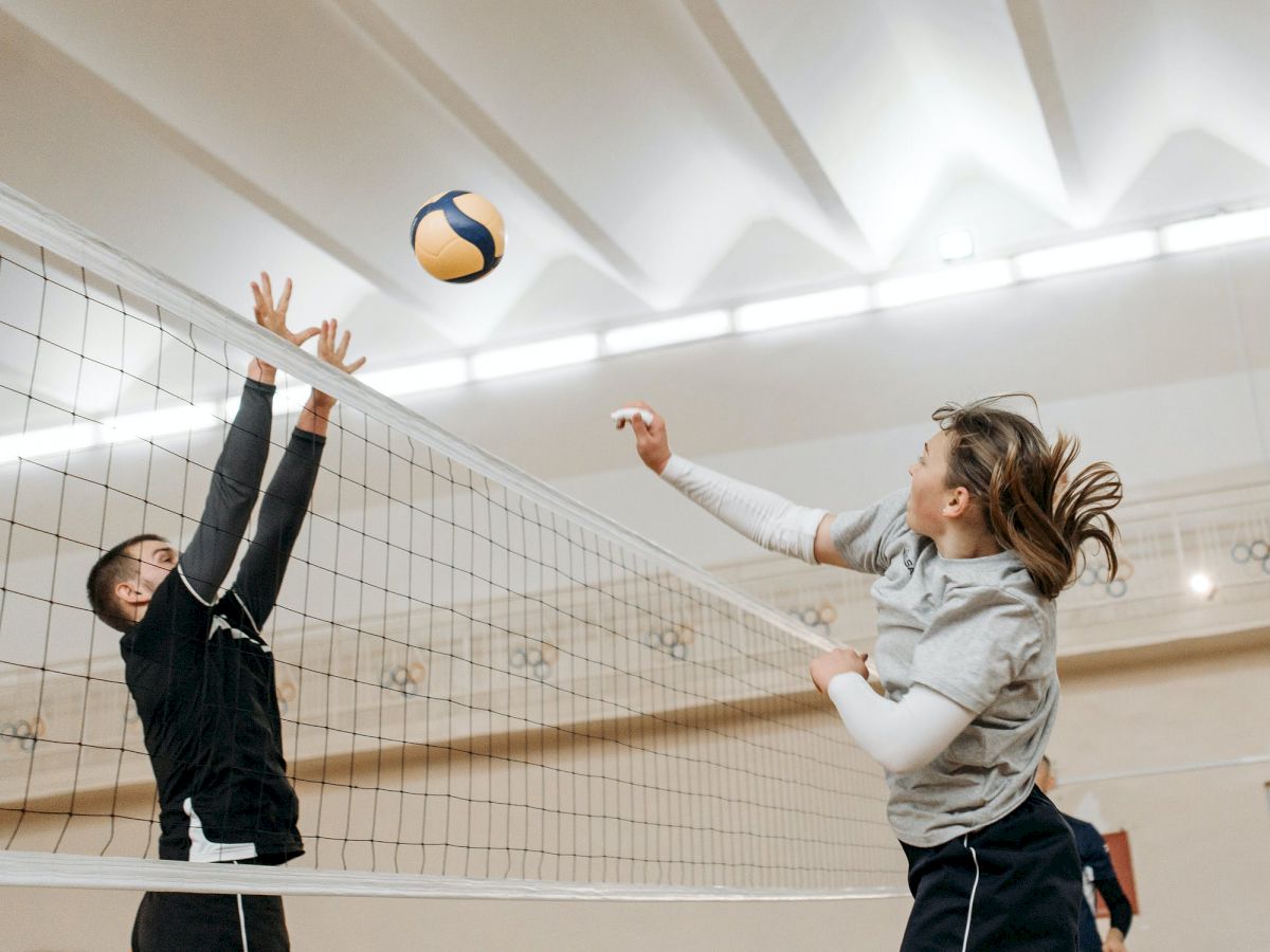 Two people are playing volleyball indoors, one jumping to spike the ball while the other attempts to block at the net.