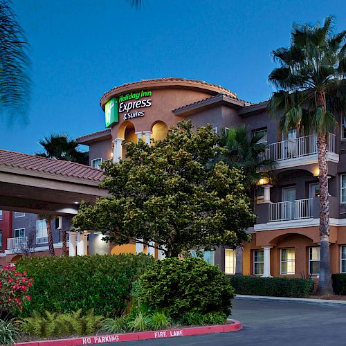 The image shows a Holiday Inn Express & Suites building with palm trees, plants, and a parking area. The hotel has multiple floors and balconies.