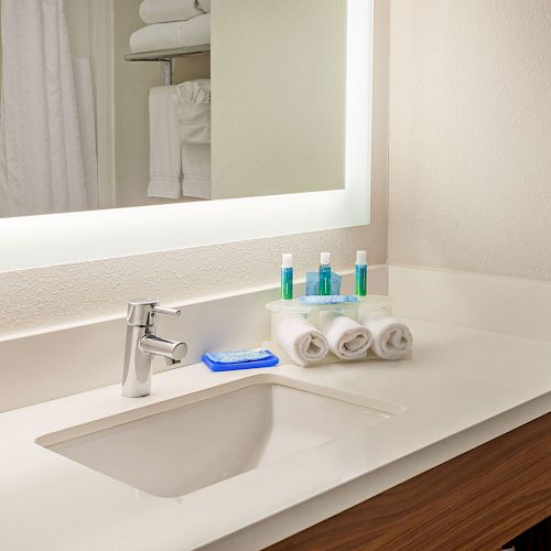 A modern bathroom countertop with a sink, faucet, neatly folded towels, and toiletries arranged next to a large illuminated mirror.