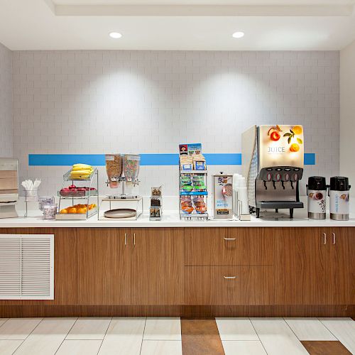 A breakfast bar area with a juice dispenser, coffee maker, cereals, and various breakfast items on a countertop in front of a gray wall.