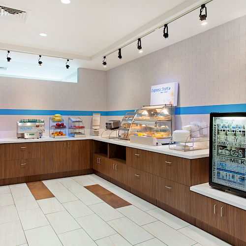 The image shows a clean, modern breakfast buffet area with various food items displayed and a milk dispenser to the right on a counter near the end.