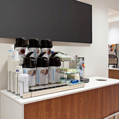 The image shows a coffee station with multiple coffee dispensers, cups, lids, condiments, and a small refrigerated display for food items.