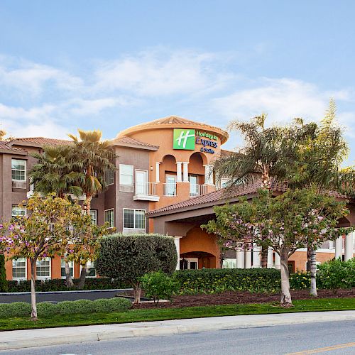 The image shows the exterior of a Holiday Inn Express hotel with a well-maintained garden and trees in front, under a partly cloudy sky.