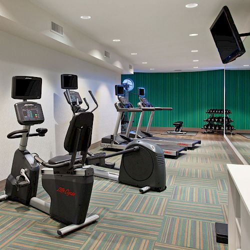 A gym with exercise bikes, treadmills, a set of dumbbells, and a TV on the wall. The floor has patterned carpeting and mirrors line one wall.