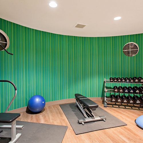 A small gym with green striped walls, exercise equipment including a bench, weights, and stability balls on a wooden floor, well-lit environment.