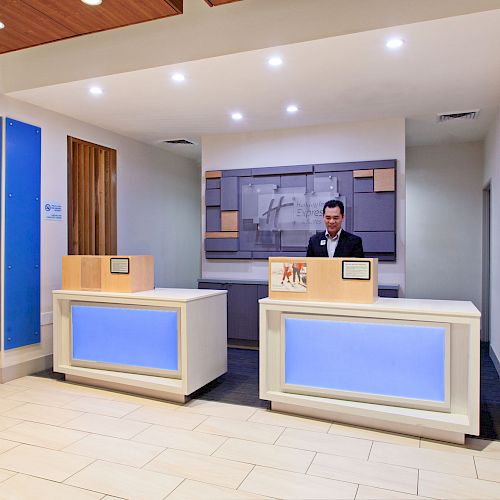 A hotel front desk area with a receptionist, illuminated blue panels, and a nearby beverage cooler.