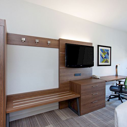 A hotel room with a wooden desk, TV, chair, wardrobe, and wall art. The room appears modern and clean, with a bed partially visible.