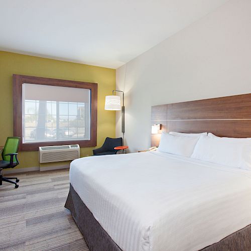 The image shows a modern hotel room with a large bed, desk, TV, green chair, and window. The decor is neat and simple.