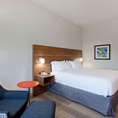 A modern, tidy hotel room with a double bed, a nightstand, a chair with a small round table, a wall lamp, and artwork on the wall.