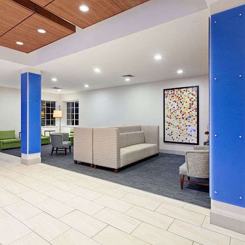 A modern lobby area with white tiled flooring, blue pillars, seating arrangements, and colorful wall art. The space is well-lit with ceiling lights.