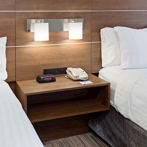 A hotel room with two beds, a wooden nightstand between them, an alarm clock, a phone, and a modern wall light fixture above the nightstand.