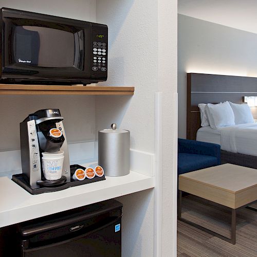 The image shows a hotel room with a bed, a microwave, a coffee maker, a mini fridge, and a table with chairs in the background.