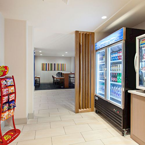 This image shows a store area with shelves of snacks, a display of candy, a refrigerated section with drinks, and a sitting area in the background.