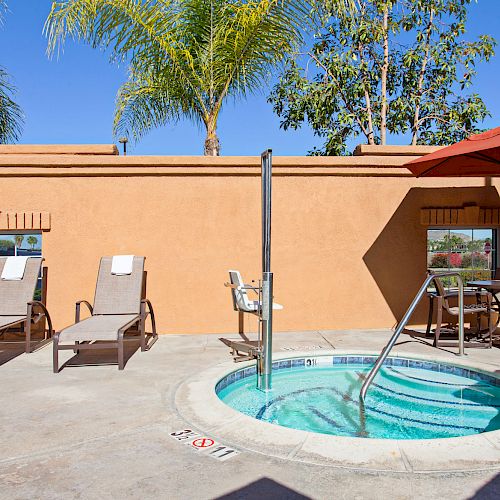 The image shows a hot tub or small pool in an outdoor area, surrounded by lounge chairs, a table with an umbrella, and palm trees in the background.