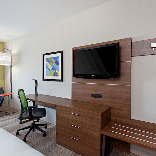 A modern hotel room with a mounted TV, desk, ergonomic chair, armchair, lamp, bed, and wall art. The room looks minimalist and organized.