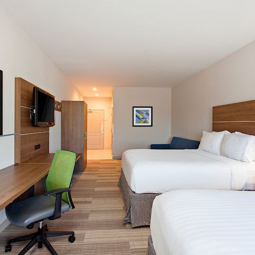 A clean, modern hotel room with two double beds, a desk, chair, wall art, TV, and bright lighting, viewed from the foot of the beds.