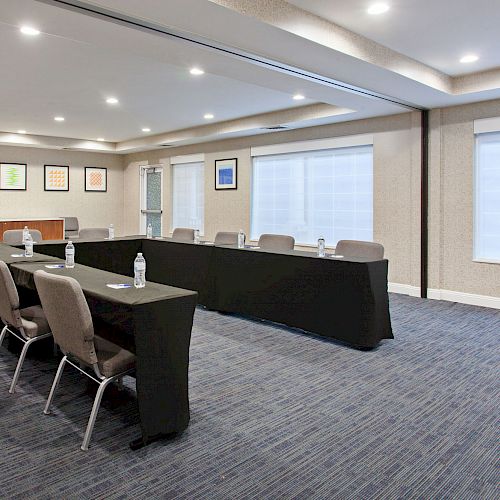 The image shows a conference room setup with tables arranged in a U-shape, chairs around them, water bottles on the tables, and framed artwork on the wall.