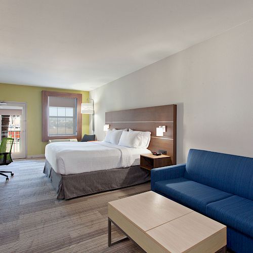 A modern hotel room with a king-sized bed, blue couch, wooden table, wall-mounted TV, a desk with a chair, and a window in the background.