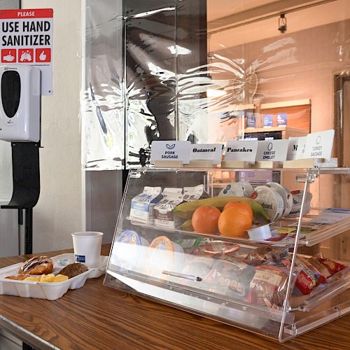 The image shows a self-serve snack bar with packaged food, fruit, and a hand sanitizer dispenser on a counter, with a hand sanitizer sign above.