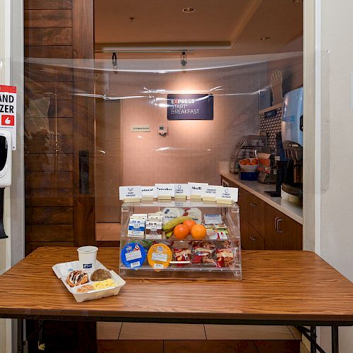 A table with breakfast items including fruit and pastries, a hand sanitizer station, and signs for breakfast hours and hand sanitization.