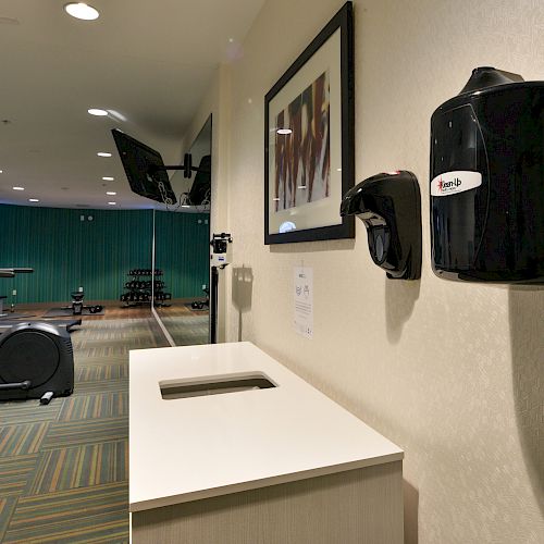 A modern gym with treadmills, an elliptical machine, a mop bucket, a towel disposal, and a hand sanitizer dispenser on the wall, with green curtains.