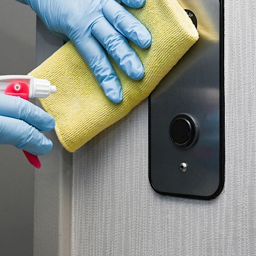 Gloved hands cleaning a door handle with a yellow cloth and disinfectant spray bottle.