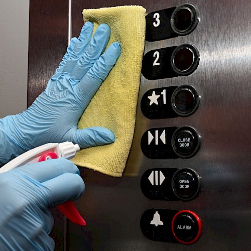 A person wearing gloves is cleaning an elevator control panel with a yellow cloth and a spray bottle.